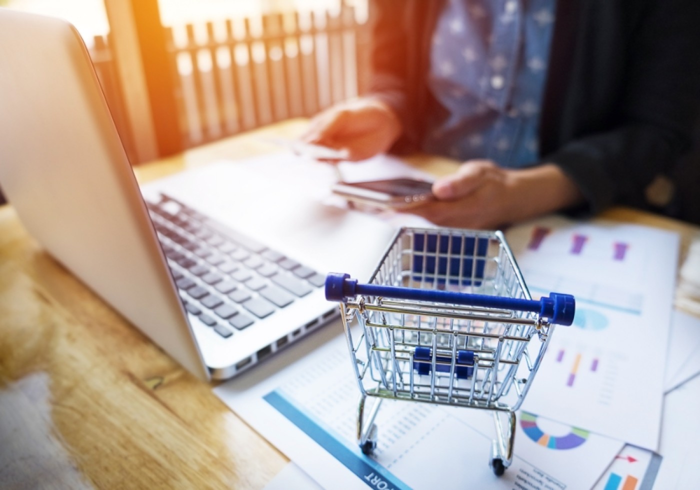 Mini shopping cart on a desk with a laptop and documents, person using a phone in the background