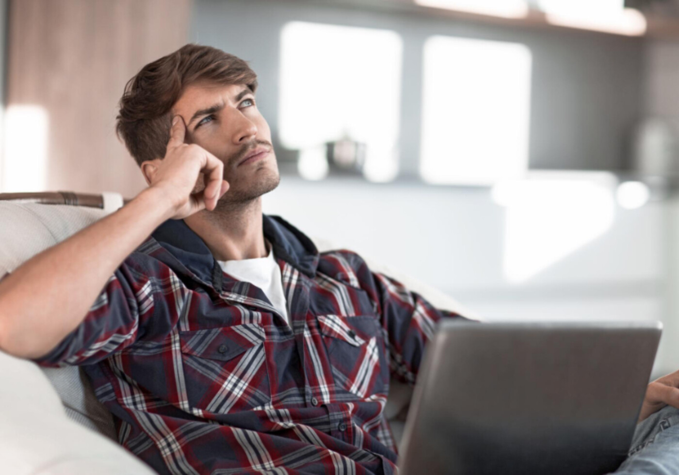 Man sat on a sofa with a laptop thinking and looking up