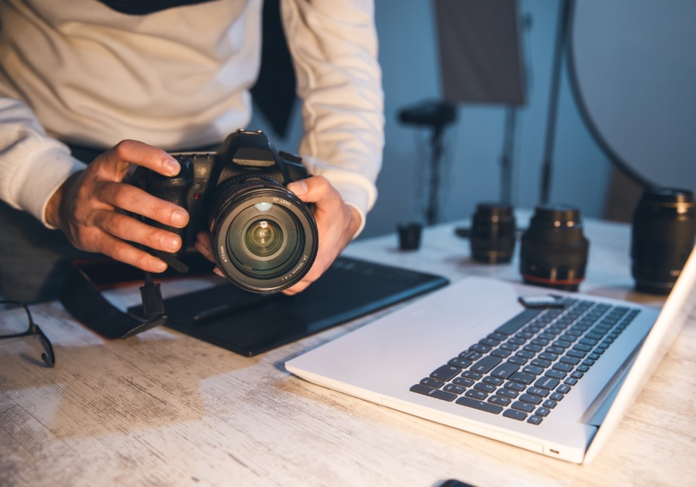 Person handling a camera on a desk with a laptop and camera lenses
