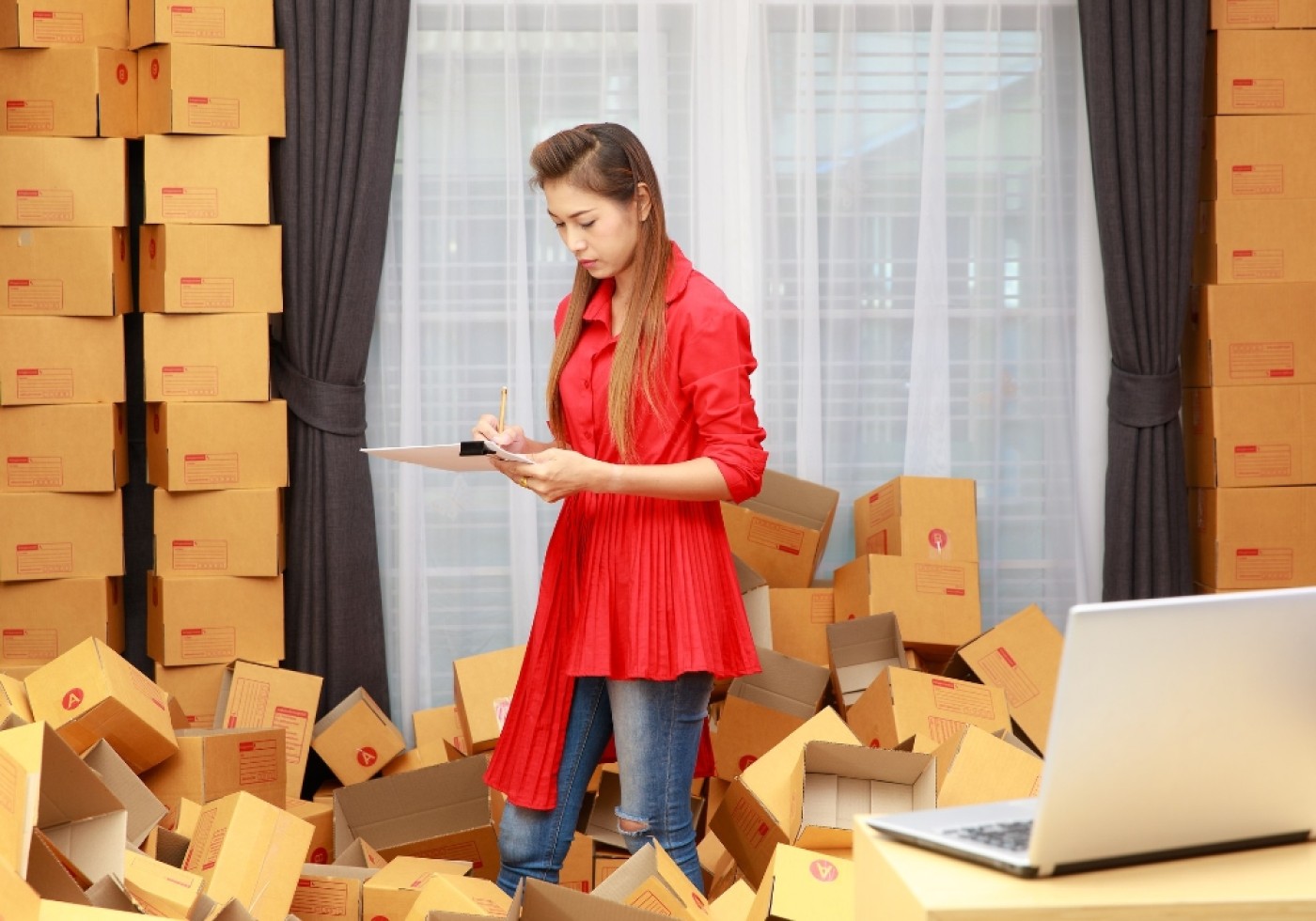 Woman checking inventory among stacks of cardboard boxes in a room.