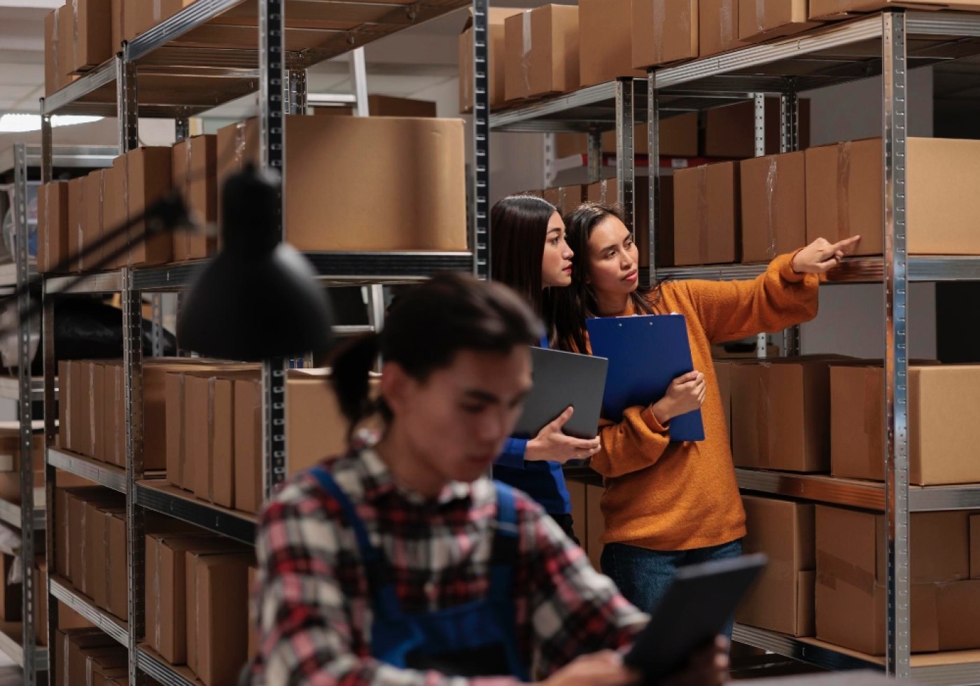 Two women discussing and pointing at shelves filled with cardboard boxes in a warehouse