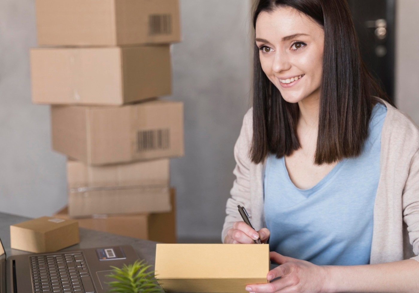 Smiling woman sitting at a desk, writing on a cardboard box with a laptop and packages in the background.