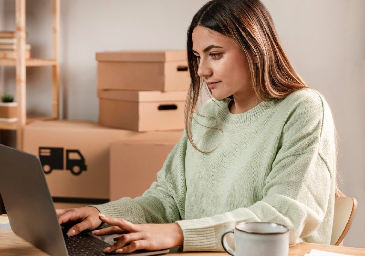 Woman in a green sweater working on a laptop with cardboard boxes in the background.