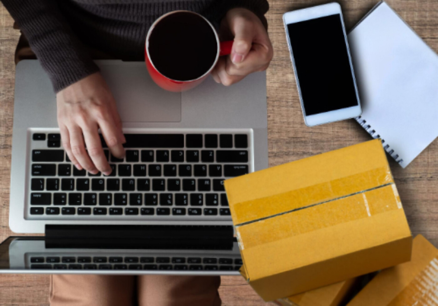 Person working on laptop with coffee mug, smartphone, notepad, and cardboard boxes.